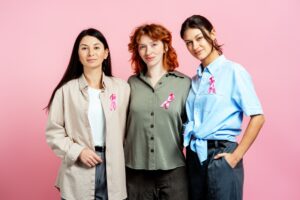 Three women standing together with pink ribbons pinned to their shirts, showing support for breast cancer awareness.