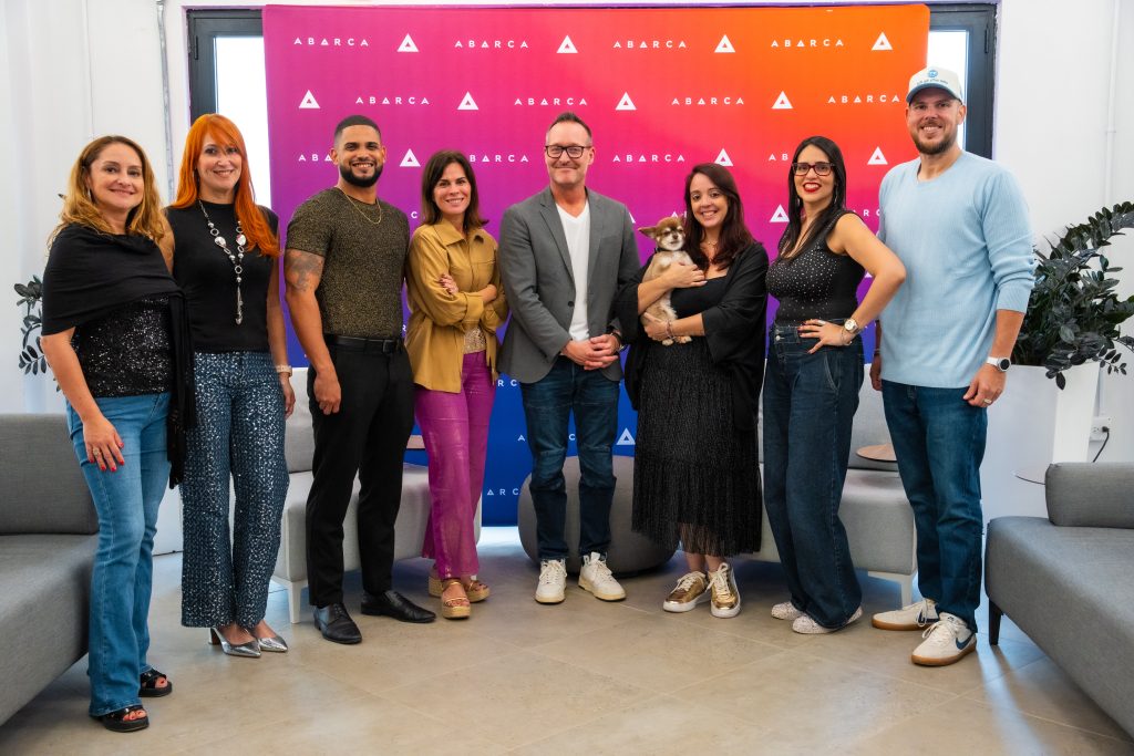 Group photo of nine Abarca team members standing and smiling in front of a colorful Abarca-branded backdrop in an office lounge, with one person holding a small dog.