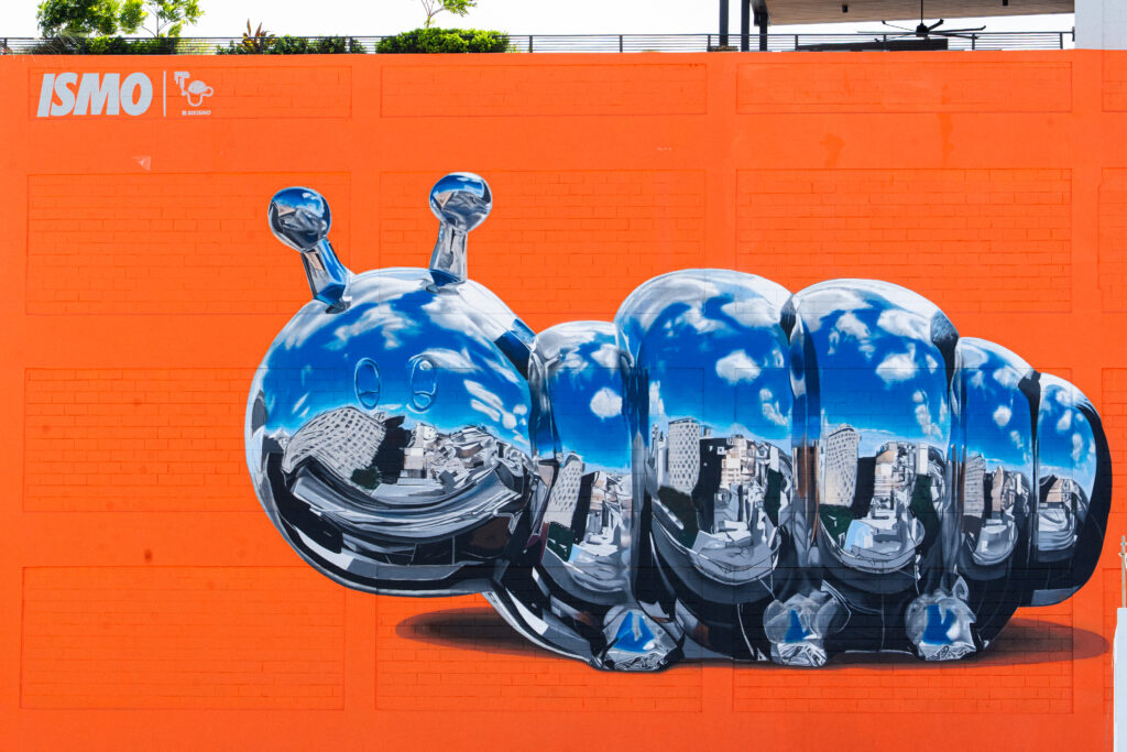 Large outdoor mural by Puerto Rican artist Bikismo on the exterior of Oasis Santurce, showing a shiny chrome caterpillar on a bright orange wall with reflections of buildings and sky.