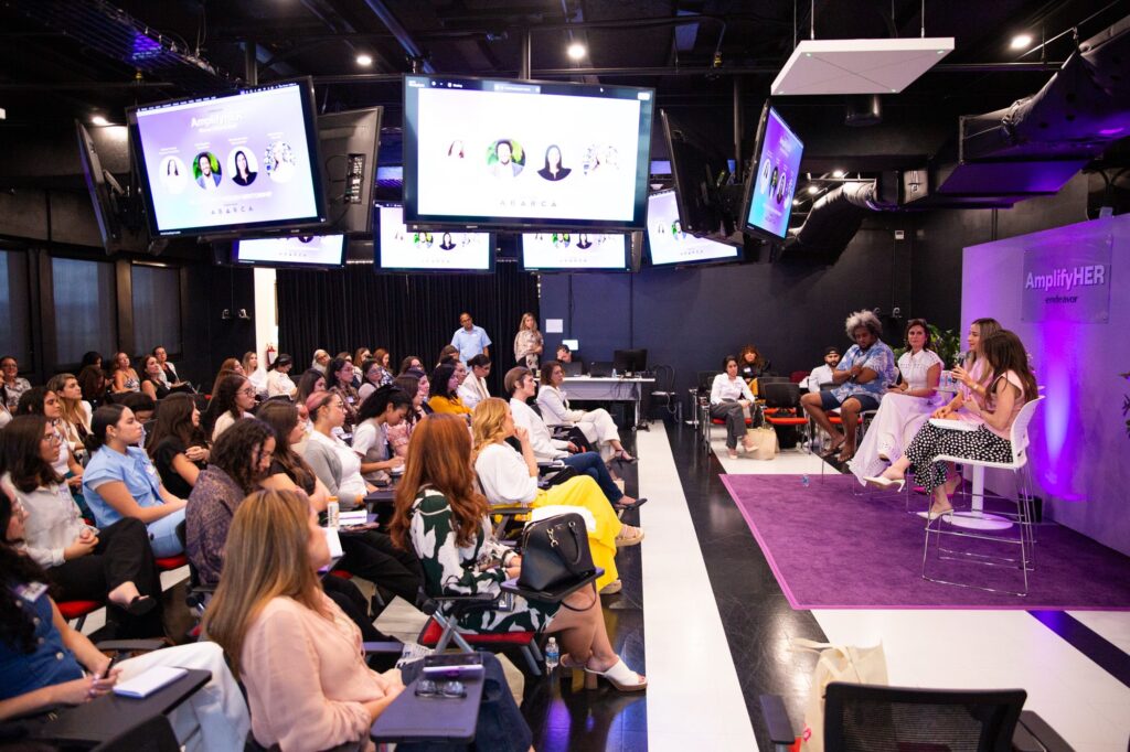 Audience seated in a modern event space listening to a panel discussion on stage at an AmplifyHER event, with speakers addressing mentorship and leadership topics while presentation screens display event visuals.