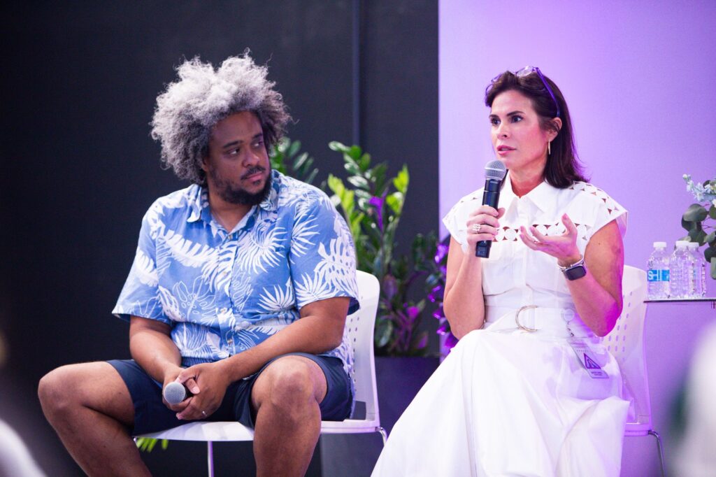 Two panelists, Mercibel Gonzalez, Executive Vice President and Chief People Officer, and Devin Baptiste of Endeavor, seated on stage during an AmplifyHER event. Mercy speaks into a microphone while Devin listens, engaged in a discussion on leadership and mentorship.
